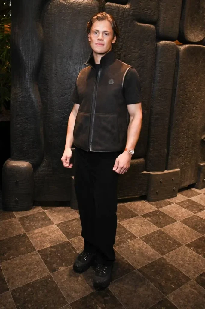 A man stands in a black leather vest, T-shirt, wide trousers and sneakers before a textured black wall