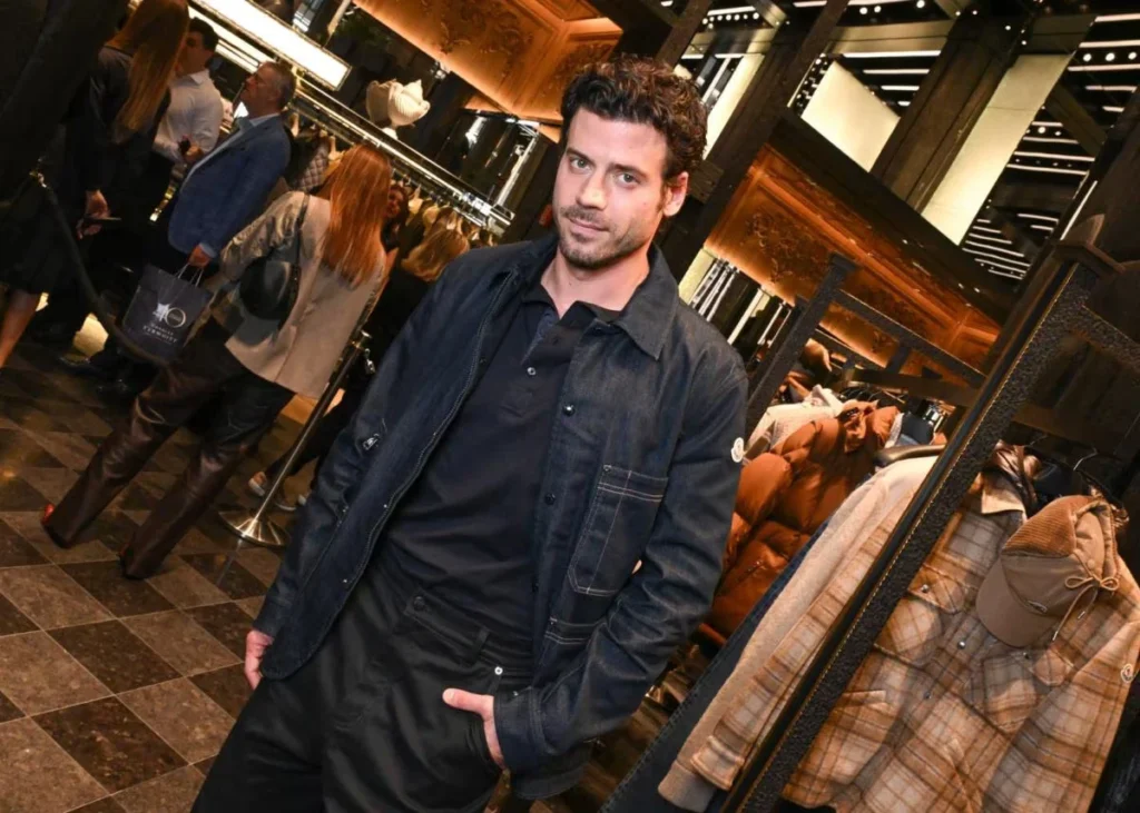 Man poses in a black jacket and polo inside a warmly lit store with puffer coats displayed nearby