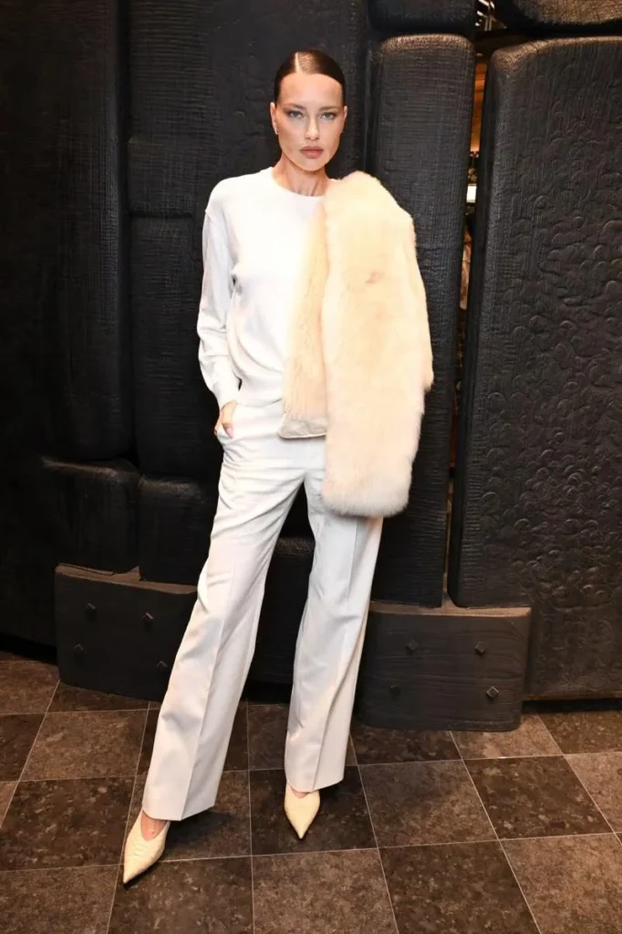 Woman in a white outfit and pointed shoes poses with a large cream fur stole against a dark textured backdrop