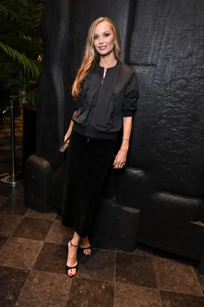 Blonde woman in a black zip-front jacket, long skirt and strappy heels posing against a black textured backdrop
