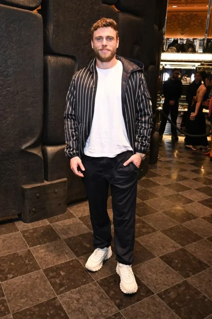 Bearded man in a black pinstriped hooded jacket, white shirt and sneakers posing inside a tiled Moncler store