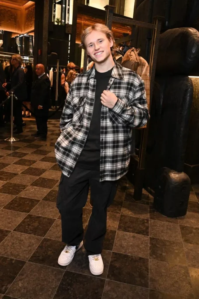Young person smiling in a black-and-white plaid overshirt, black clothes and white sneakers inside a warmly lit boutique