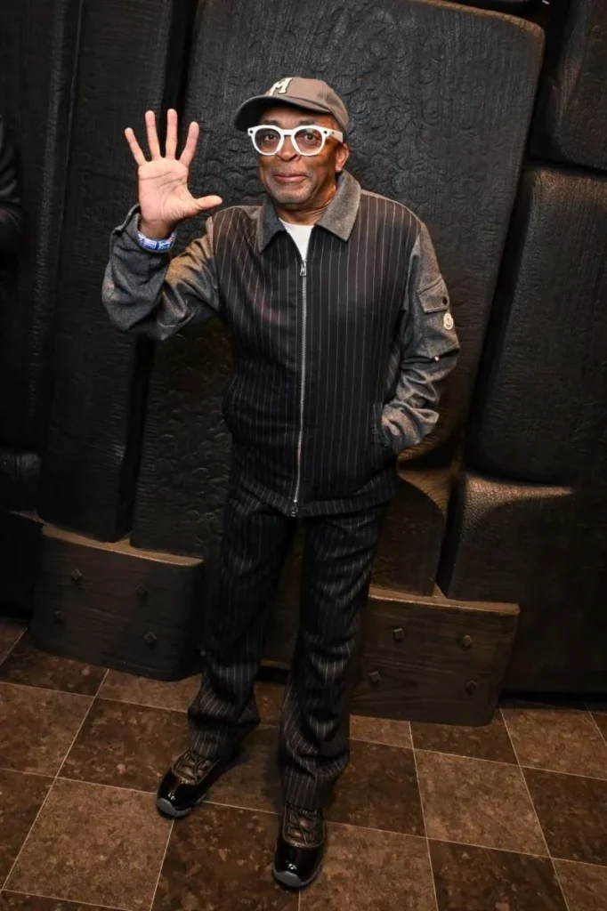 Spike Lee waves in a pinstriped jacket and pants, white glasses, and a brown cap against a dark padded wall