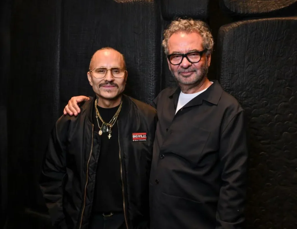 Two men pose together against a black textured backdrop; one wears glasses, layered necklaces, and a black jacket with a red