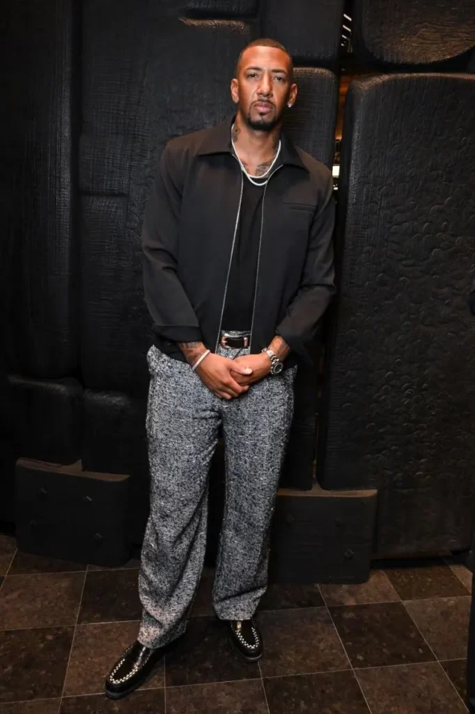 Man stands with hands clasped, wearing a black jacket and shirt, gray textured trousers, and black loafers against a dark
