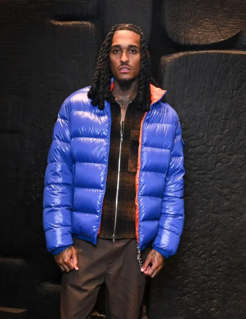 Man with long braids wearing a glossy blue puffer jacket over brown clothing, standing against a textured black wall