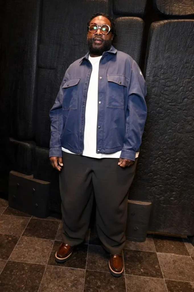 A man wearing round glasses, a blue overshirt, white shirt, black trousers, and brown shoes stands against a black wall