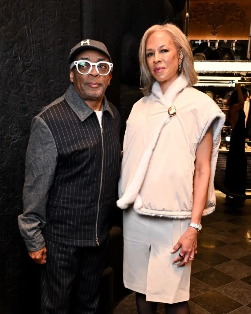 Spike Lee and a woman pose inside a boutique; he wears white glasses and a pinstriped jacket, she wears a cream fur-trimmed