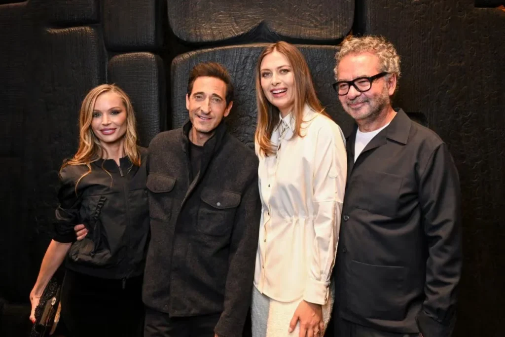 Anne Hathaway and three guests pose together against a black textured wall; Hathaway wears a white belted shirt dress
