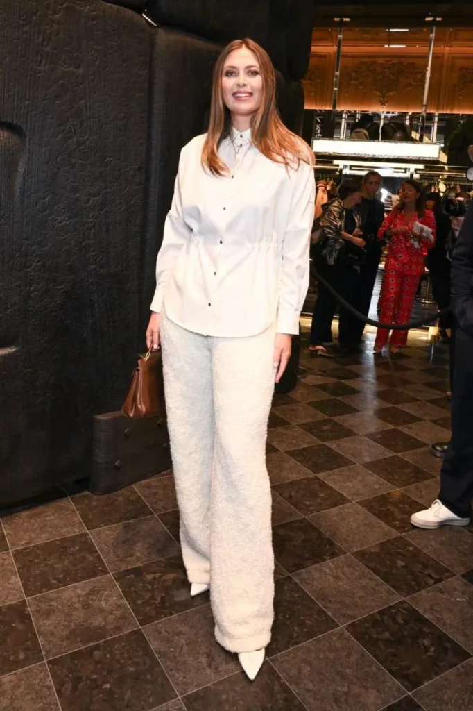 Anne Hathaway poses in a white button-down and textured wide-leg trousers, holding a brown handbag at a Moncler event