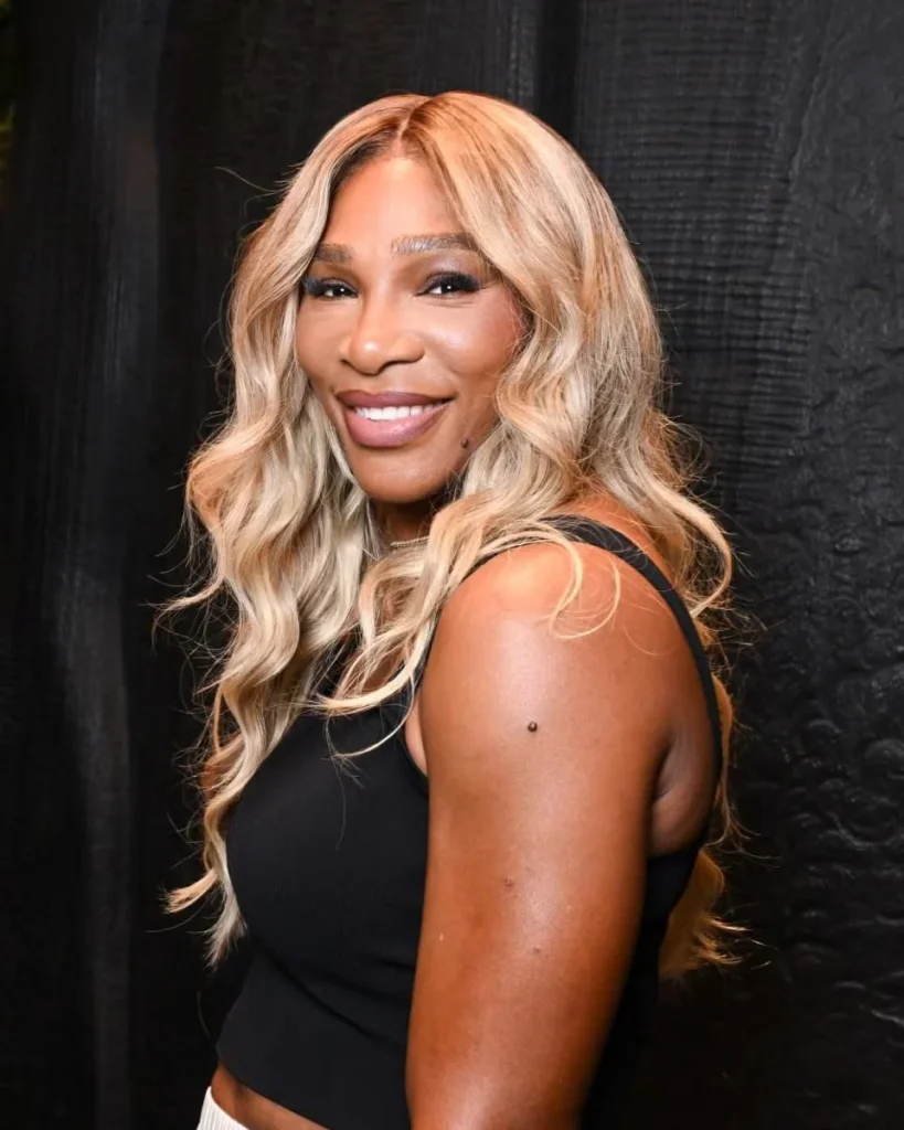 Serena Williams smiles over her shoulder in a black sleeveless top against a dark textured backdrop