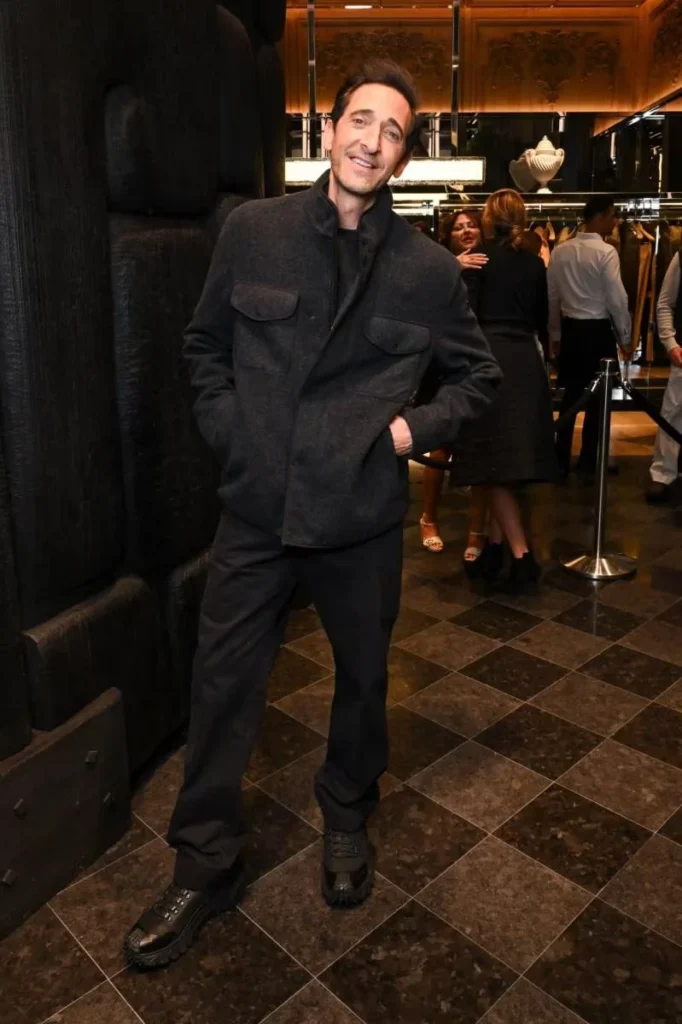 Adrien Brody poses in a dark jacket and trousers inside a warmly lit luxury store with guests behind him