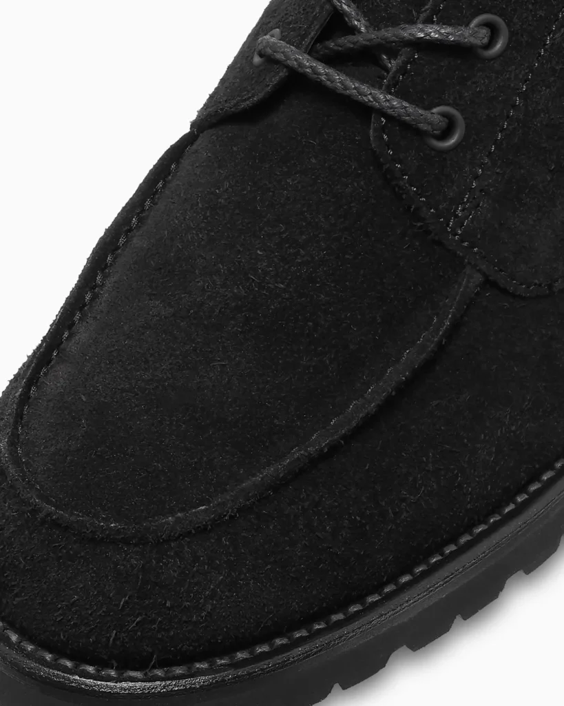 Close-up of a black suede moccasin-style shoe with braided laces, metal eyelets, stitched toe, and lugged sole