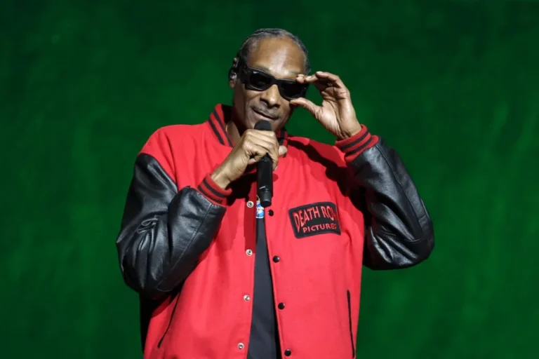 Snoop Dogg holds a microphone and adjusts black sunglasses, wearing a red varsity jacket with black leather sleeves onstage