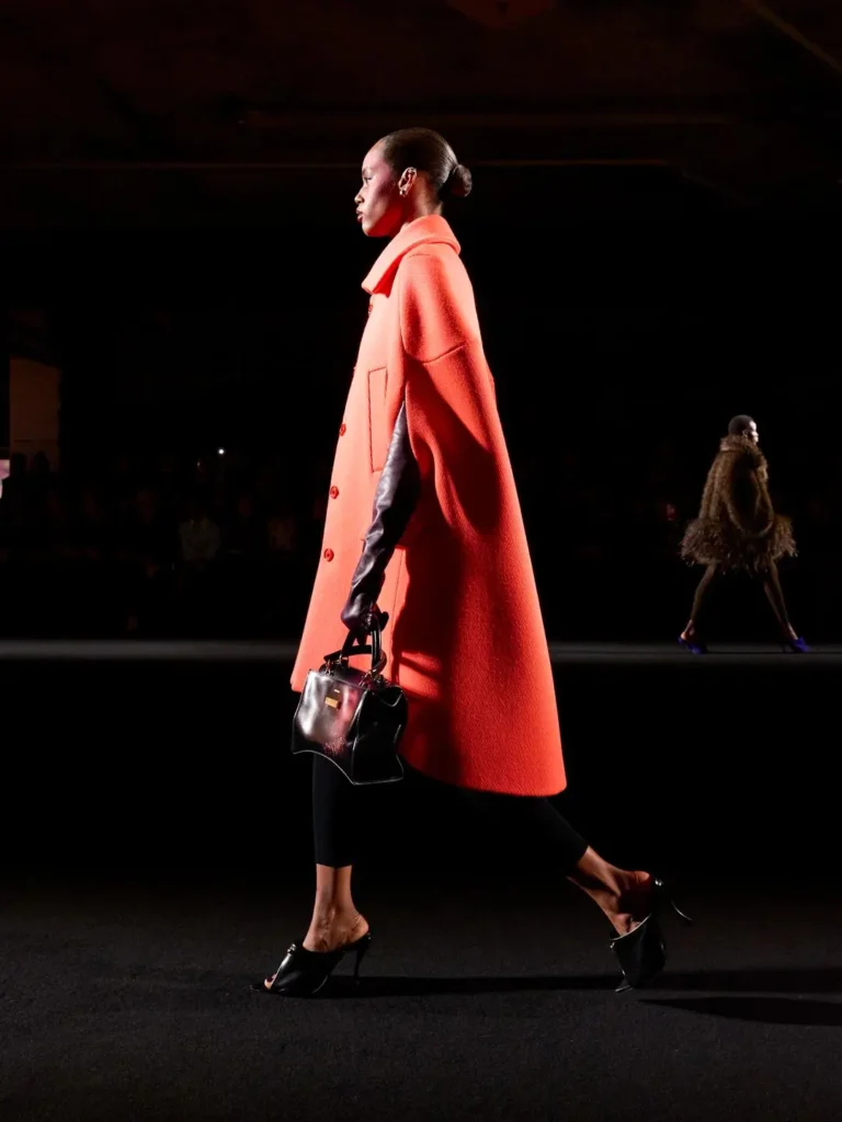 Model walks runway in a coral oversized coat, black leggings, black handbag and mule heels under spotlights
