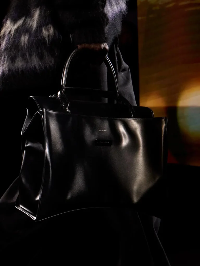 Person in a black fur-textured coat carries a large structured black leather handbag against a dark background