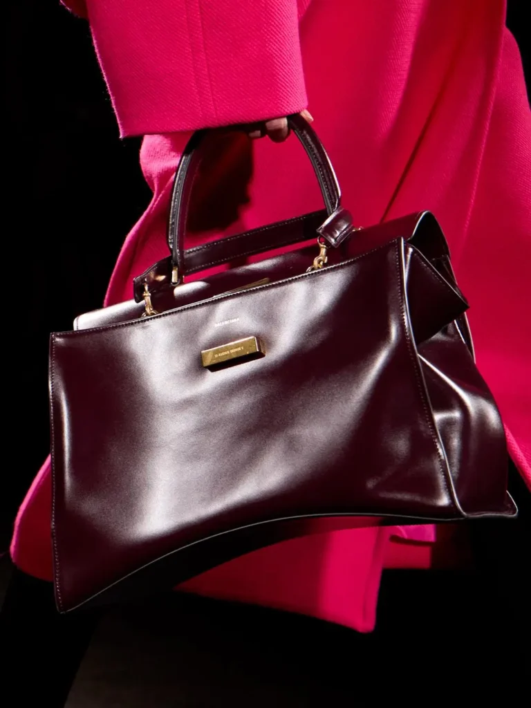Close-up of a burgundy leather handbag with structured handles against a bright pink outfit and black background