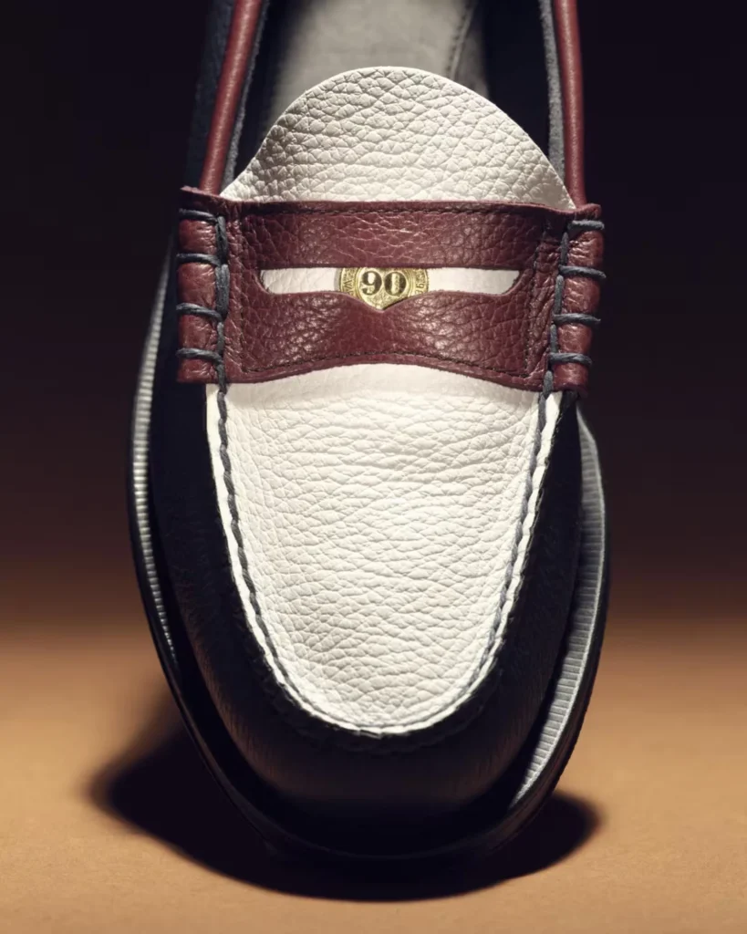 Close-up of a burgundy and white leather loafer with braided lacing and a gold 90th-anniversary emblem, on a wooden surface
