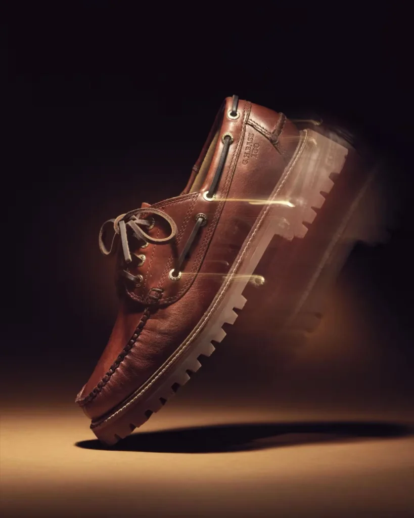 Brown leather lug-sole shoe tilted on its toe, with black laces and metal eyelets, lit against a dark background