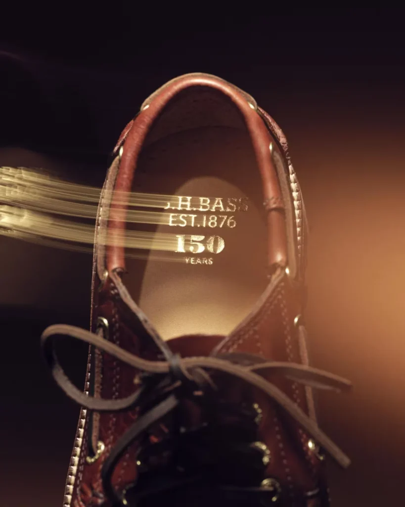 Close-up of a burgundy leather shoe with laces, showing “J.H. Bass Est. 1876 150 Years” on the insole amid warm light streaks