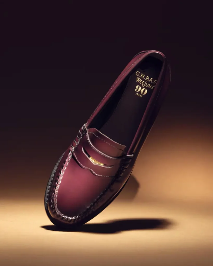 Burgundy leather penny loafer tilted on its toe, casting a shadow on a warm tan surface against a dark background