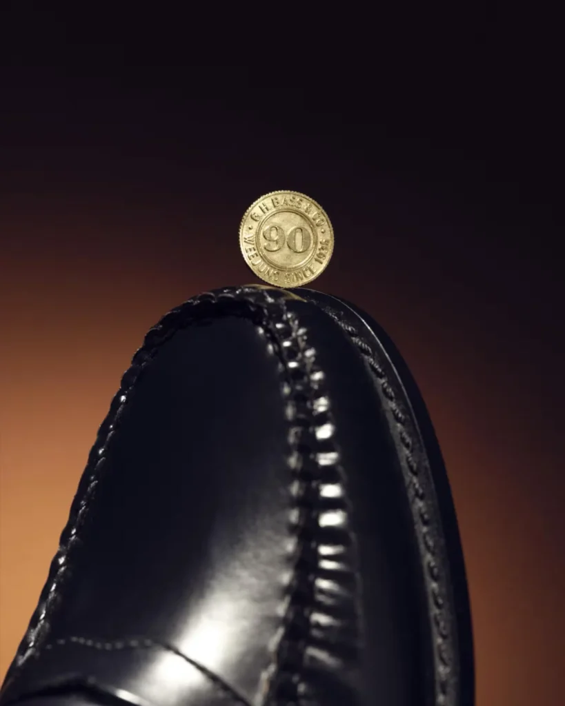 Close-up of a black leather loafer balanced beneath a gold 90th-anniversary coin against a dark brown background