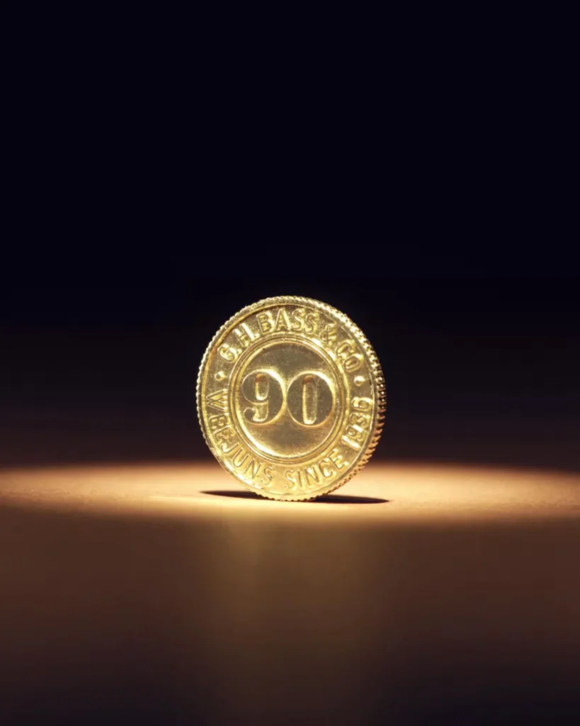 Gold coin marked “G.H. Bass & Co.” and “90,” standing on its edge on a warmly lit surface against a dark background