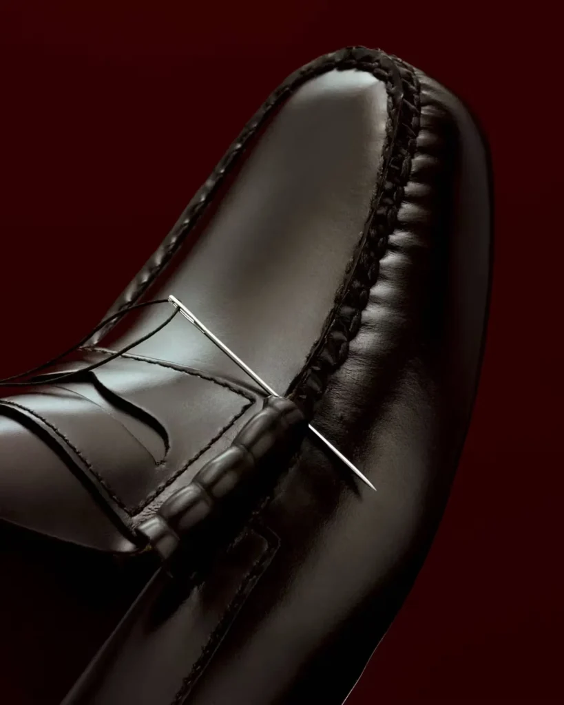 Close-up of a black leather loafer with braided trim and a silver needle-like ornament against a dark red background
