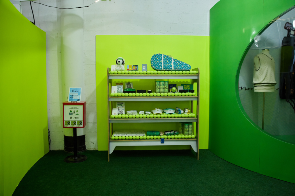 Shelves display Snoopy tennis-themed merchandise lined with tennis balls in a neon green exhibition space