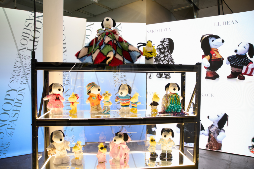 Snoopy and Woodstock plush figures in colorful designer outfits displayed on illuminated glass shelves beside digital screens