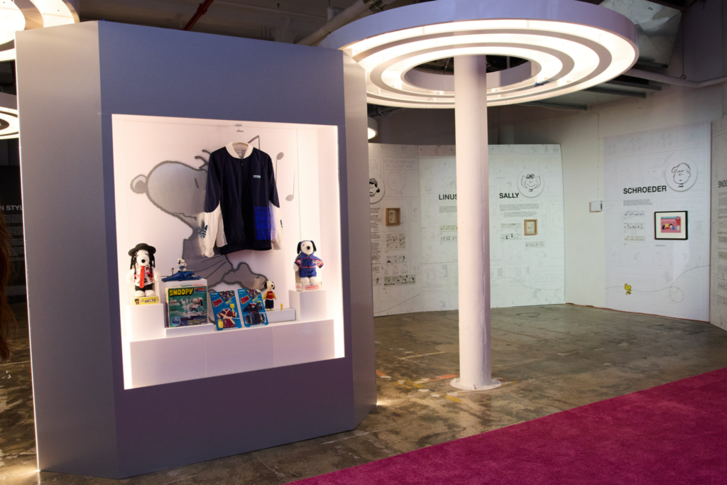 Peanuts exhibition with Snoopy toys and clothing in a lit display case, comic panels on the walls, and a magenta carpet