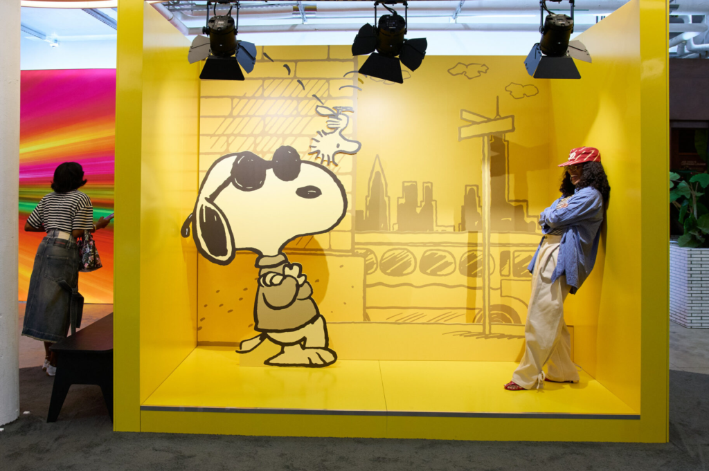 Snoopy cutout stands in a yellow New York-themed display beside a woman in a red cap, blue shirt, and cream pants