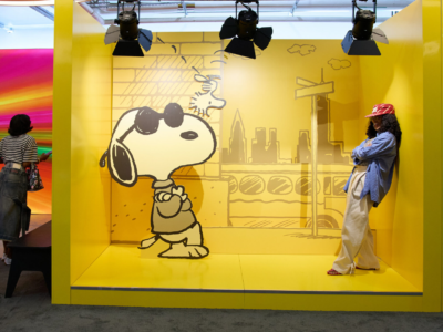 Snoopy cutout stands in a yellow New York-themed display beside a woman in a red cap, blue shirt, and cream pants