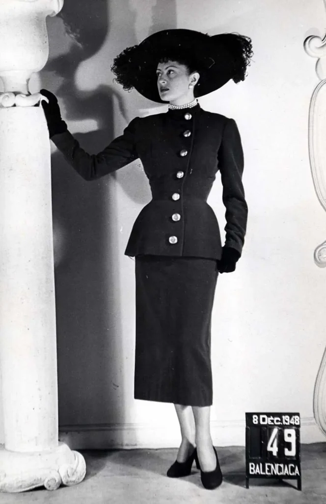 Woman in a black Balenciaga suit, jeweled choker and broad hat poses beside a column; 1948 placard at right