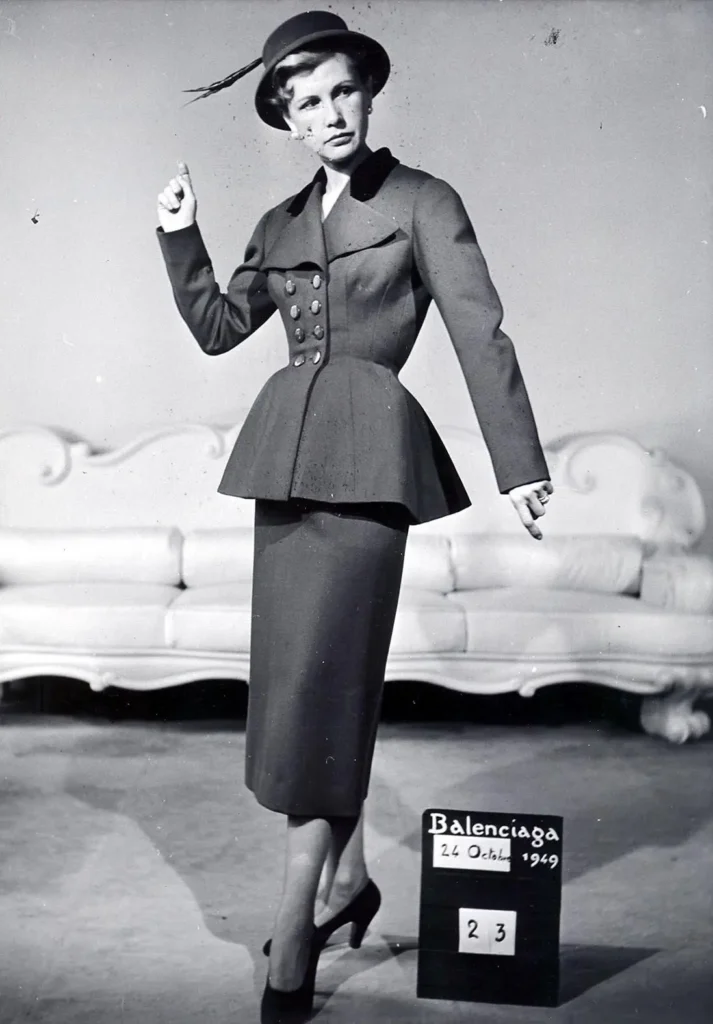 Model poses in a structured peplum jacket and knee-length skirt with a feathered hat, beside a Balenciaga 1949 display