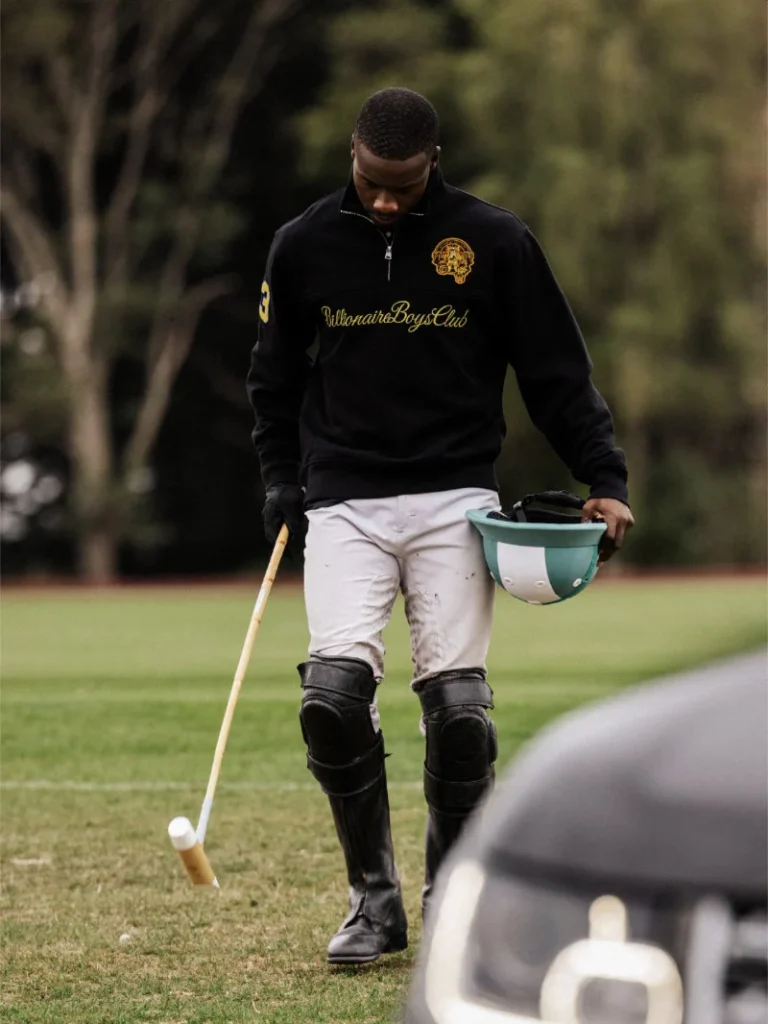 Polo player in black sweatshirt and protective gear walks across a field carrying a helmet and mallet