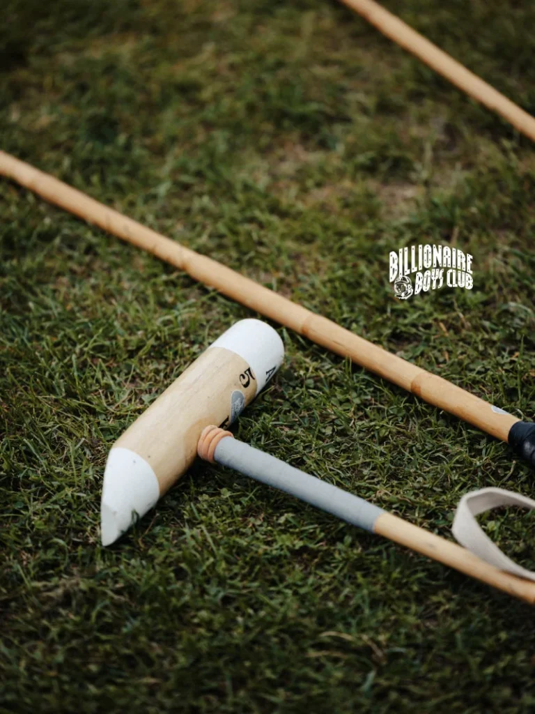 Wooden polo mallets with white heads lie crossed on grass, with a Billionaire Boys Club logo visible near the center right