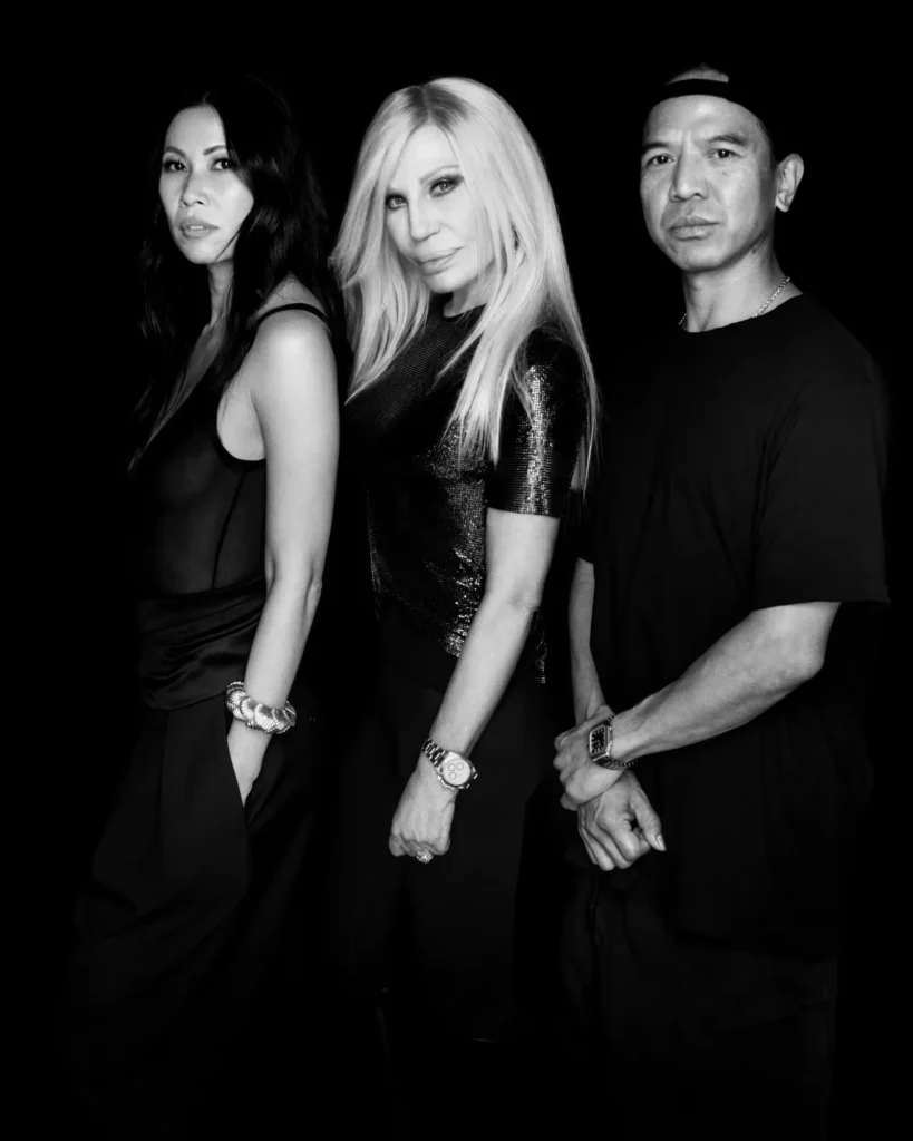 Three pose against a black background; the center woman wears a shimmering black top and watch