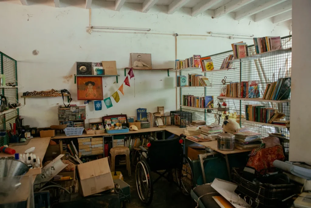 Cluttered room with bookshelves, stacked papers, a wheelchair, framed portraits, and colorful flags affixed to white walls