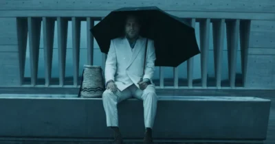 Johnny Depp sits on a dock in a white suit, holding a large black umbrella beside a woven basket