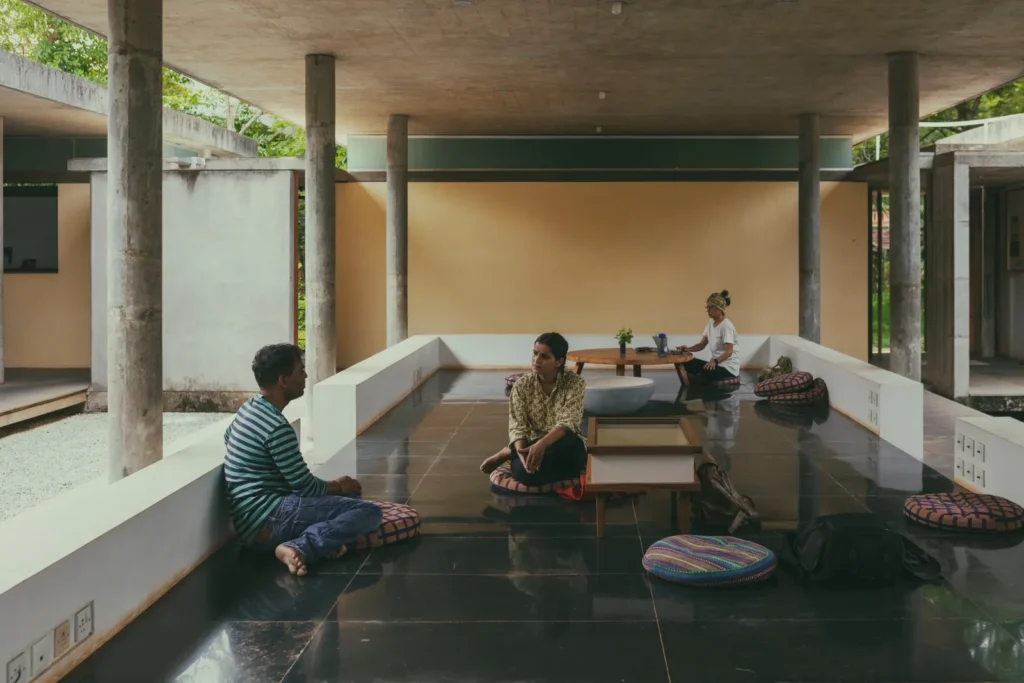 Three people sit on colorful floor cushions in an open concrete pavilion with columns, dark tile floors, and tropical