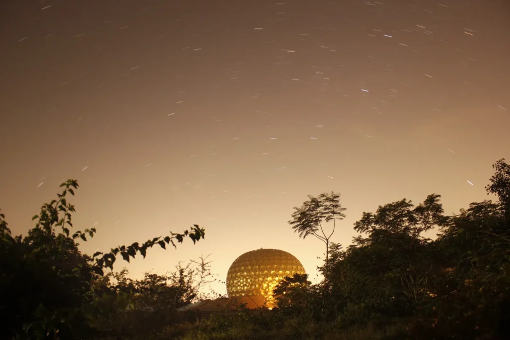 Golden spherical building glows among trees beneath a sky streaked with star trails at dusk