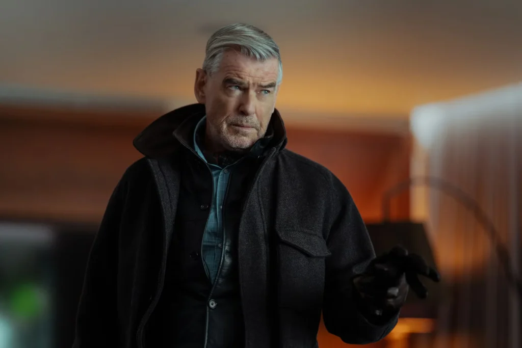 Pierce Brosnan wears a dark coat and gloves, raising one hand against a warm blurred interior backdrop