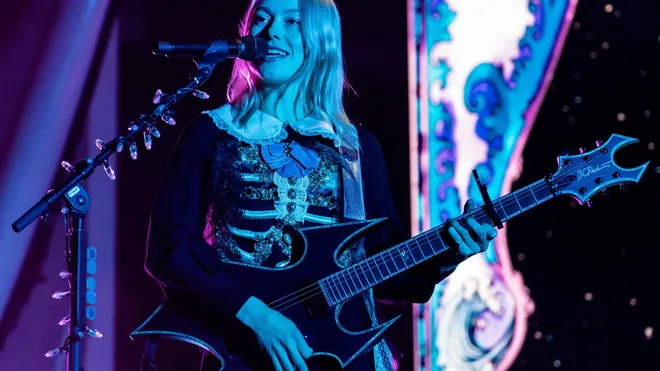 Phoebe Bridgers sings into a microphone while playing a black electric guitar on a blue-lit stage
