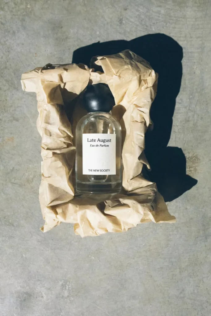 Glass perfume bottle label “Late August Eau de Parfum” nestled in crumpled beige paper on a concrete surface