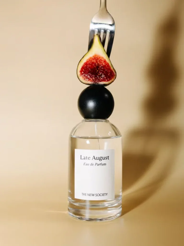 Clear glass perfume bottle label “Late August” beneath a black sphere, fig half, and fork against a beige background