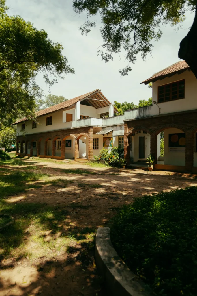 White stucco buildings with red-tile roofs and brick arches surround a shaded, tree-lined courtyard in Auroville