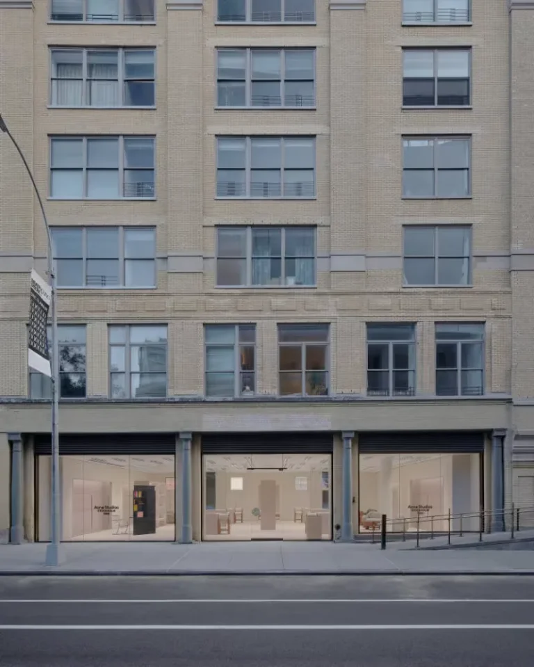 Acne Studios storefront with three glass display windows beneath a beige brick apartment building on a city street