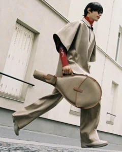 Person strides through a street carry a tan guitar case, wearing a beige cape and trousers with a red sleeve marked 927