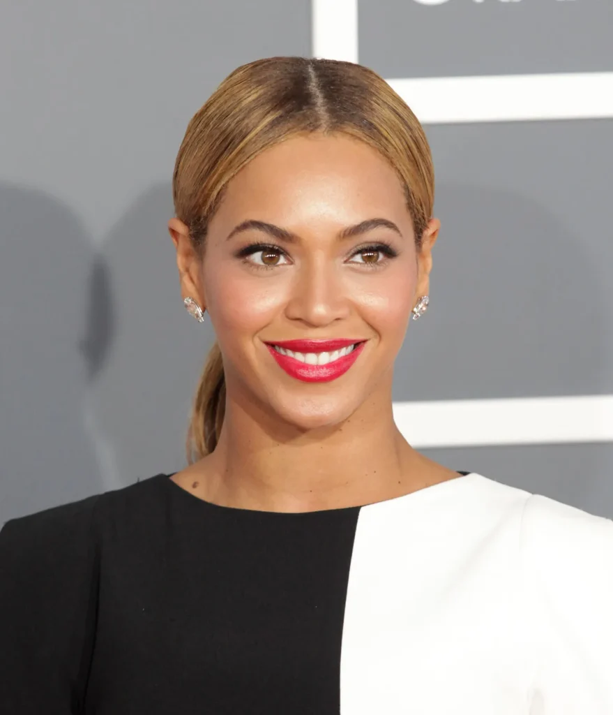 Beyoncé smiles in a close-up portrait wearing a black-and-white dress, red lipstick, diamond earrings, and a sleek ponytail